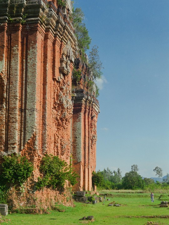 A person walks across a grassy field next to a large, weathered brick structure under a clear sky.