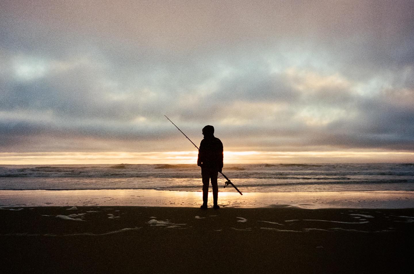 A man stands on a beach holding a fishing rod at sunset.