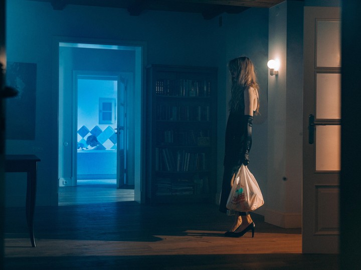 A woman in a black dress and gloves stands in a dimly lit room, holding a plastic bag, with a bright blue bathroom visible through a doorway in the background.