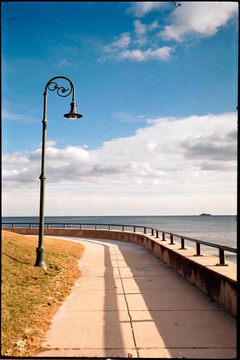 A curved path with a lamppost runs alongside a body of water under a partly cloudy sky.
