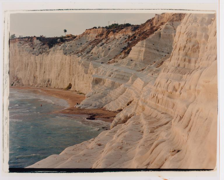 A sandy beach is nestled at the base of towering white cliffs, with a single palm tree visible at the top of the cliff.