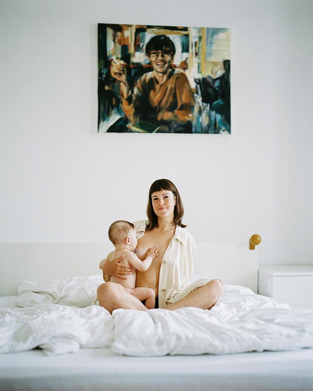 A woman sits on a bed holding a baby, with a painting of a man on the wall behind her.