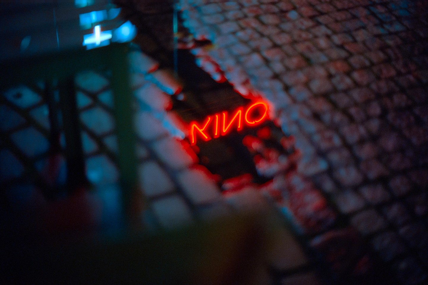 A red neon sign that reads "KINO" is reflected in a puddle on a cobblestone street at night.