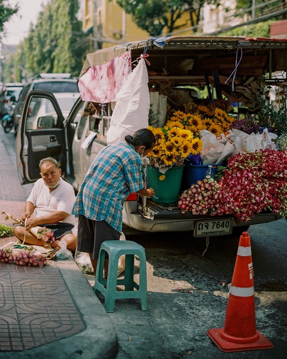 Two people arrange flowers on a street next to a truck filled with sunflowers and red blossoms.