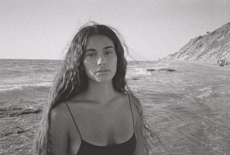 A woman with long, wavy hair stands in the ocean with a cliff and beach in the background.