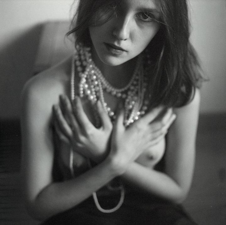 A woman with long hair and multiple strands of pearls around her neck covers her chest with her hands.
