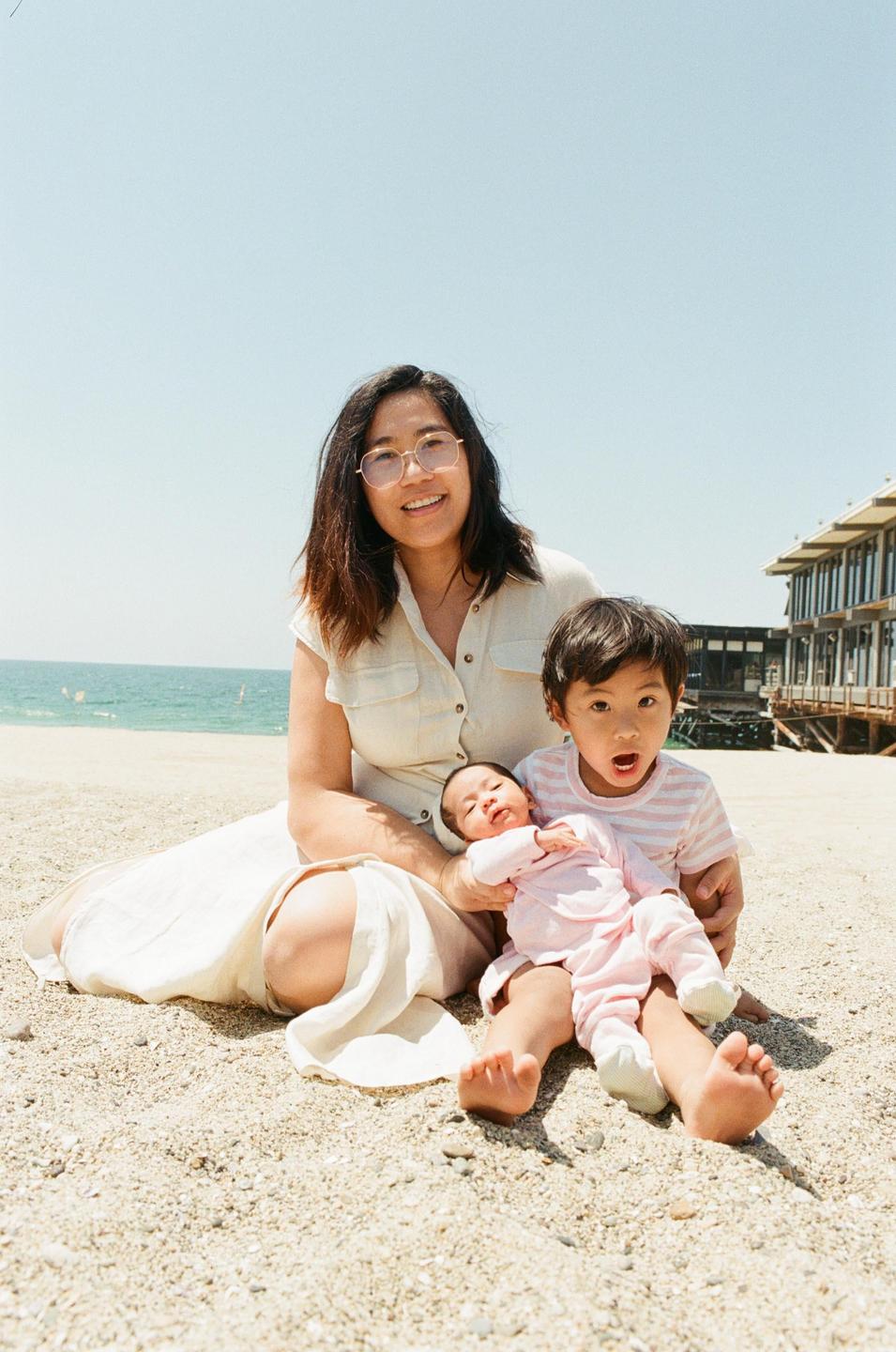A woman sits on a sandy beach with a boy and a baby, with the ocean and a building in the background.