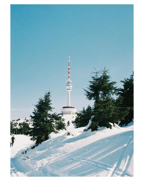 A tall white and red tower stands on a snowy hill surrounded by evergreen trees under a clear blue sky.