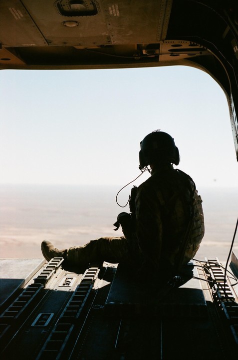 A person in military gear sits in the open doorway of a helicopter, looking out at a hazy landscape.