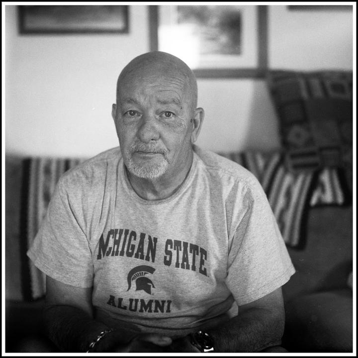 A man with a goatee and a Michigan State Alumni t-shirt sits in a room with framed pictures on the wall.
