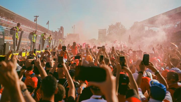 A large crowd of people with their arms raised, many holding up cell phones, are enveloped in red smoke at a race track.