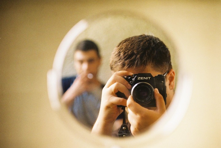 A man holds a camera to his face, reflected in a mirror, with another man out of focus in the background.