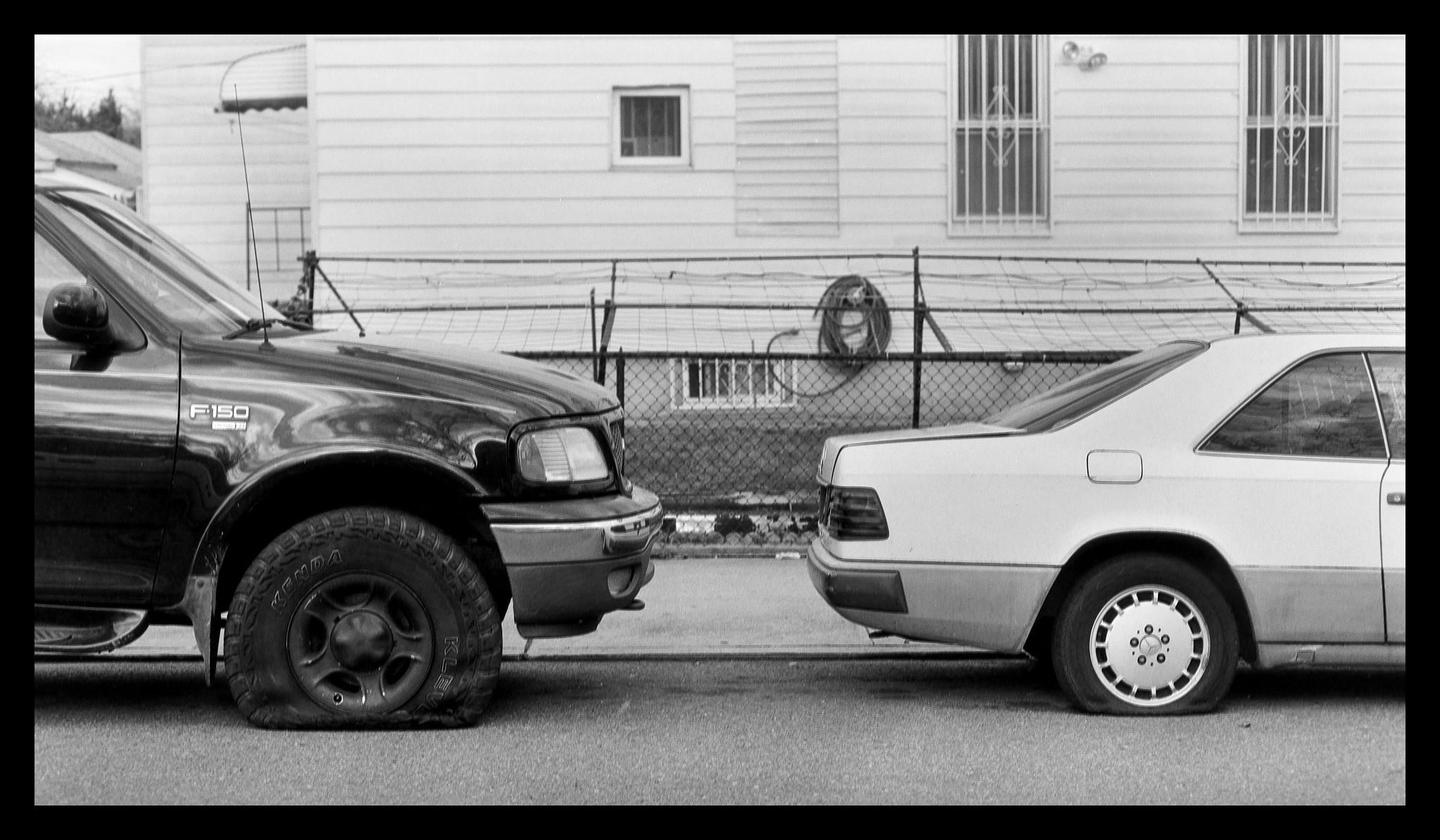 Two cars with flat tires are parked on a street in front of a house with a fence.