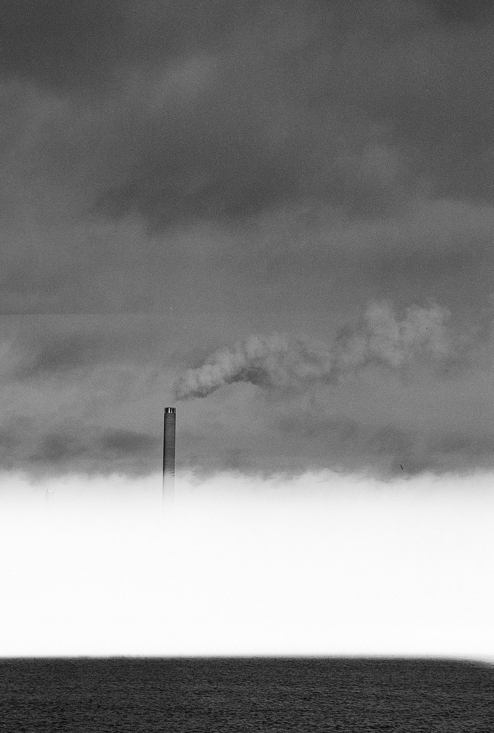A tall chimney emits smoke into a cloudy sky above a body of water.