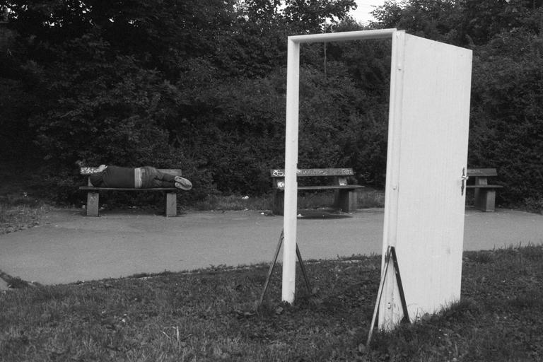 A person is sleeping on a park bench next to a white door frame and a white door standing upright in the grass.