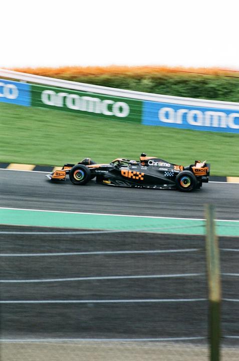 A black and orange race car speeds along a track with a green and blue banner in the background.