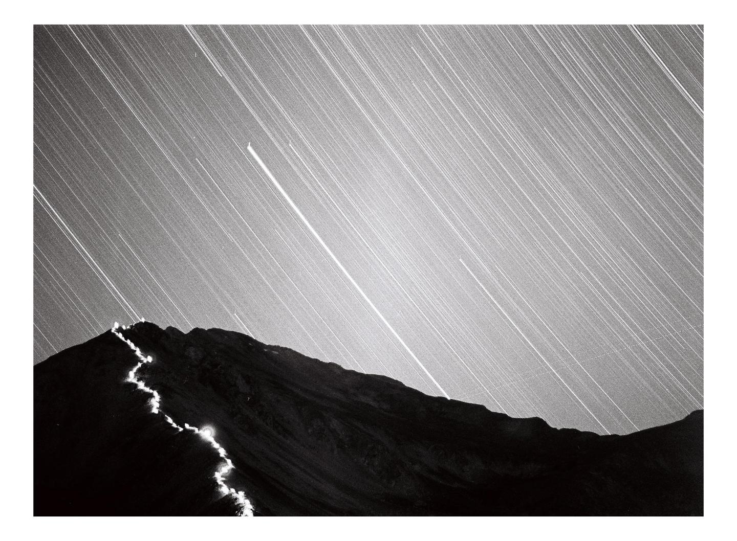 A mountain peak is illuminated by a winding trail of light under a sky filled with star trails.