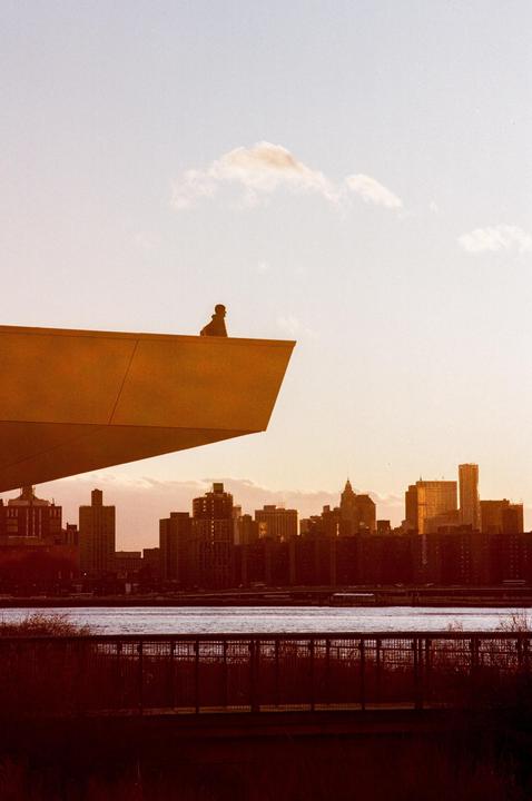 A person sits on the edge of a modern building overlooking a city skyline and river at golden hour.