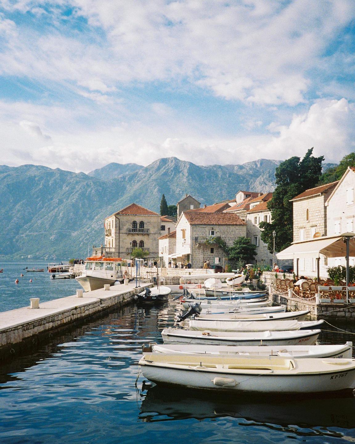 A row of small boats is docked along a stone pier in a bay with buildings and mountains in the background.