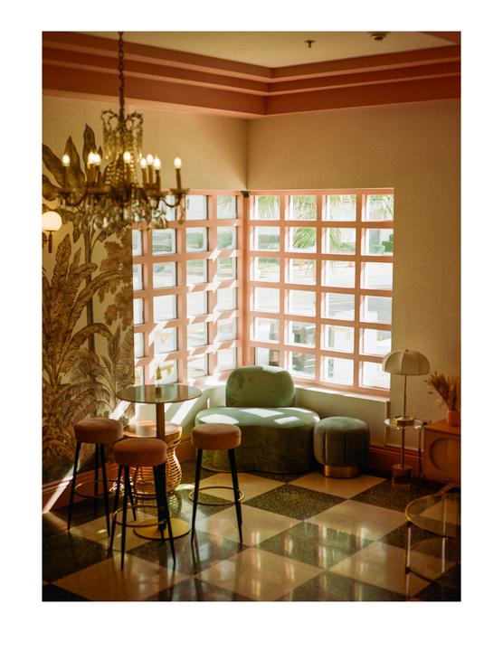 A room with a checkered floor features a small table with stools, a green velvet chair, and a large window with pink frames.