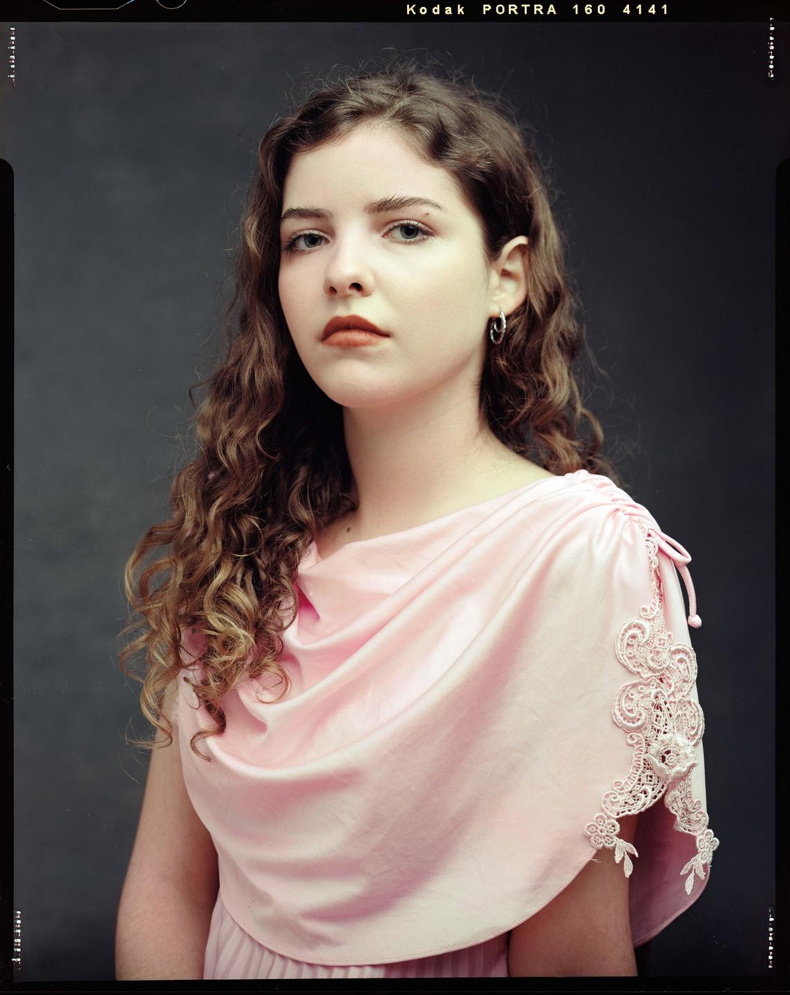 A young woman with curly hair wears a pink dress with lace details.