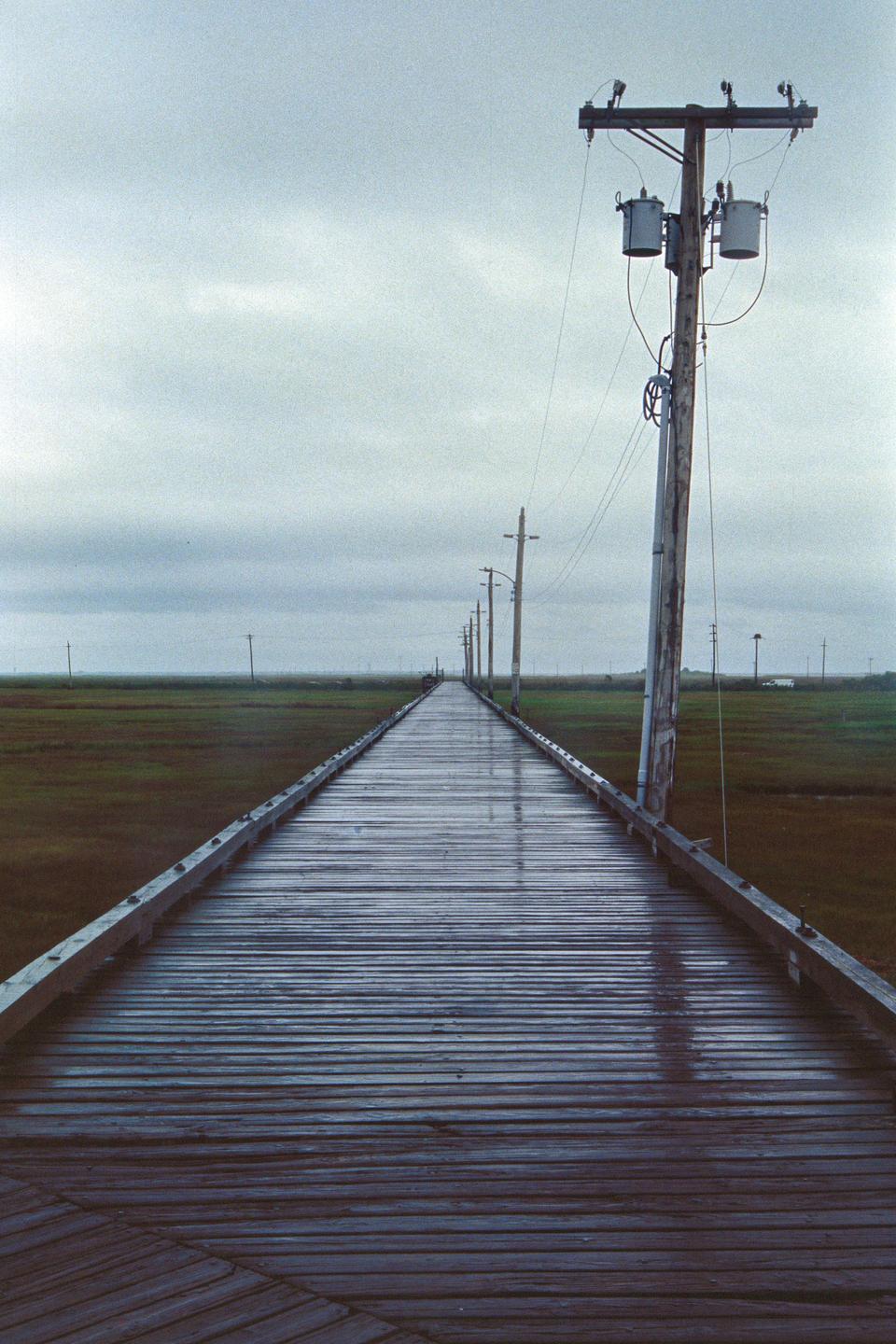 A long wooden causeway stretches into the distance, lined with utility poles and surrounded by marshland under a cloudy sky.