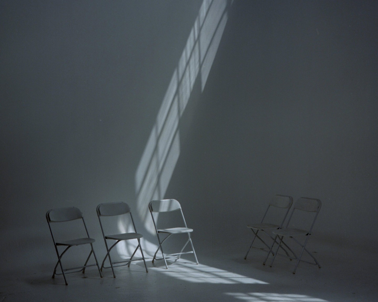 Five folding chairs are arranged in a room with a strong diagonal shadow cast across the wall and floor.