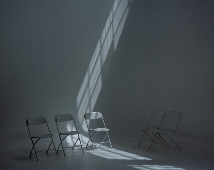 Five folding chairs are arranged in a room with a strong diagonal shadow cast across the wall and floor.