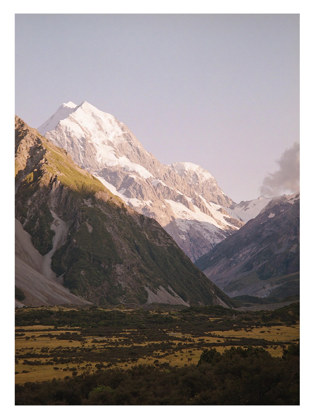 A snow-capped mountain peak rises above a valley with green and brown vegetation under a clear sky.