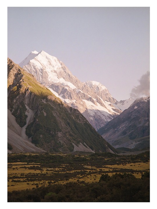 A snow-capped mountain peak rises above a valley with green and brown vegetation under a clear sky.