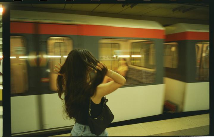 A woman stands on a subway platform as a train passes by, with the word "JUNGFERNS" visible on the train.