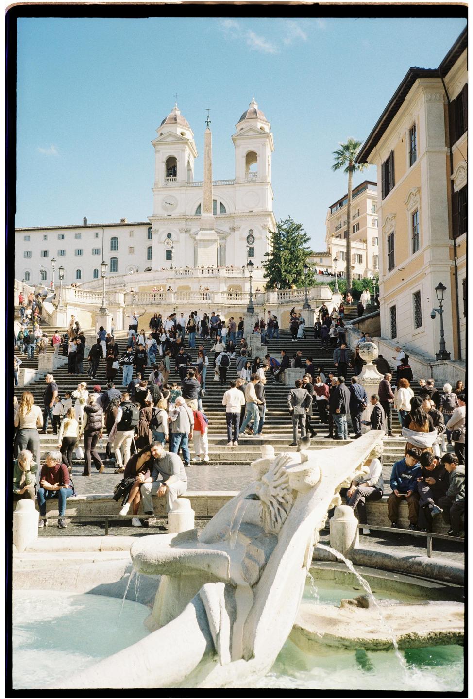 A large crowd of people are gathered on the Spanish Steps in Rome, with a fountain in the foreground and a church in the background.