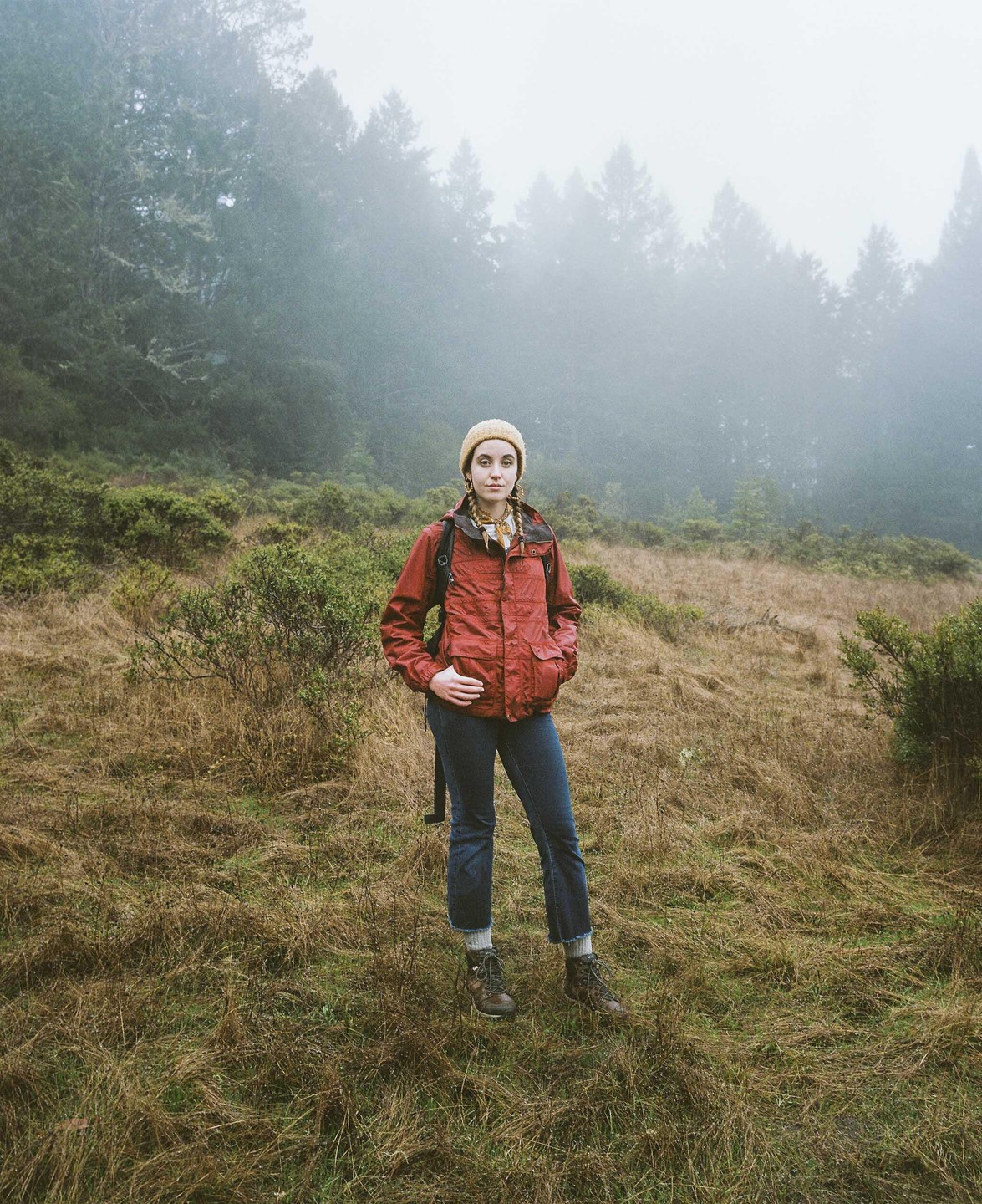 A woman stands in a field with trees and fog in the background.