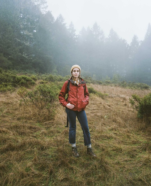 A woman stands in a field with trees and fog in the background.