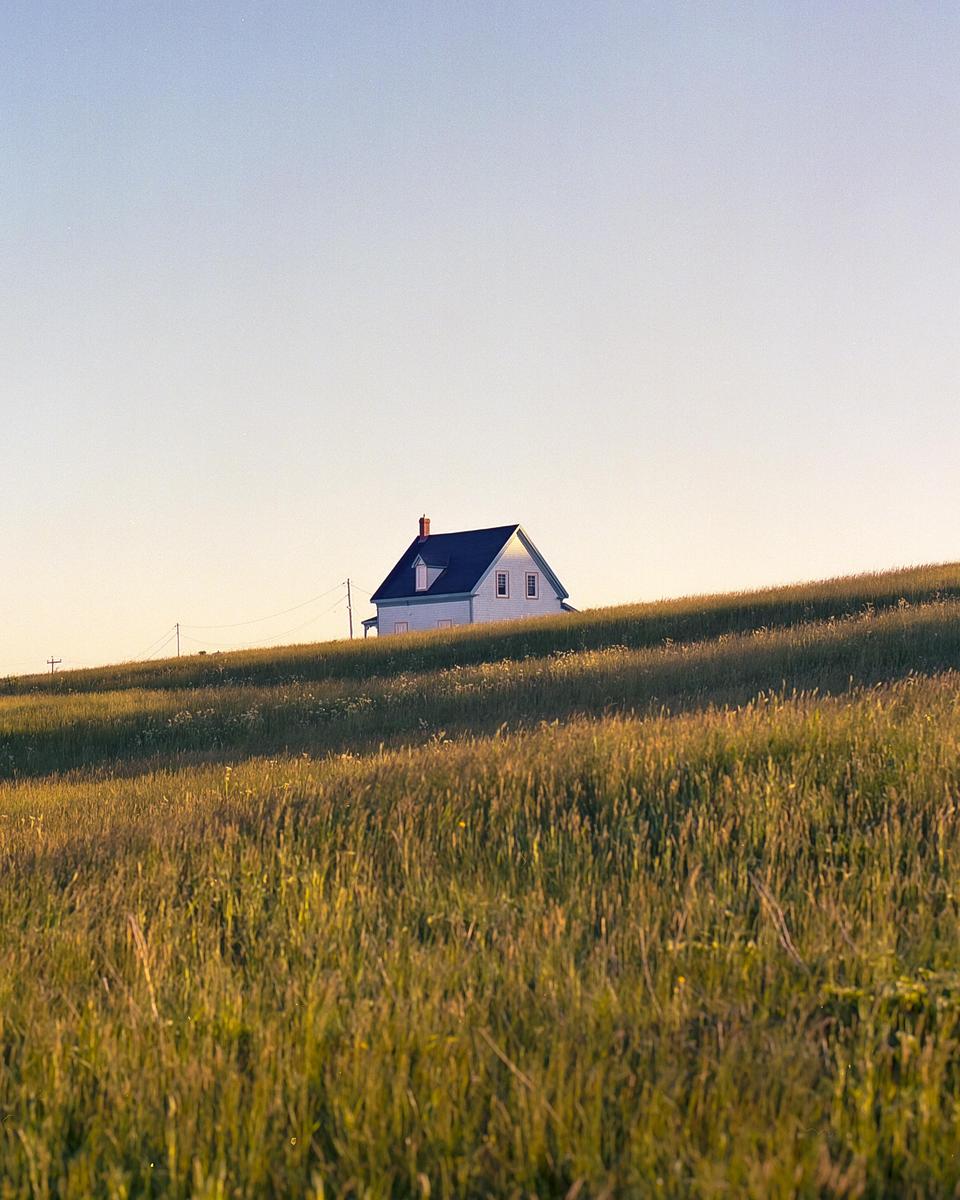 A white house with a dark roof sits on a grassy hill under a clear sky.