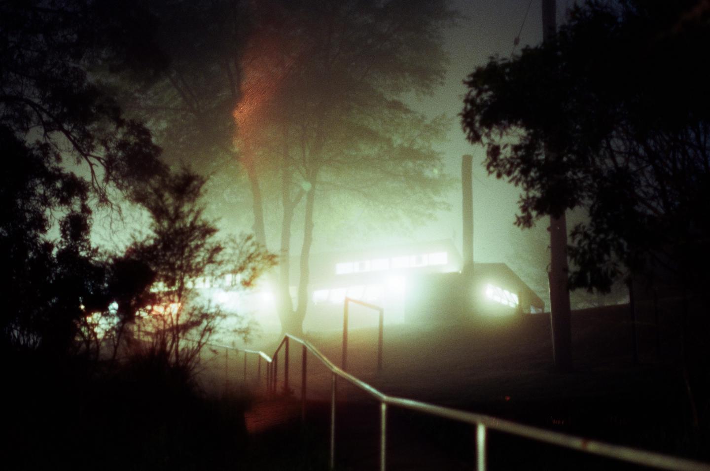 A foggy night scene shows a building with illuminated windows and a handrail leading down a path, surrounded by trees.