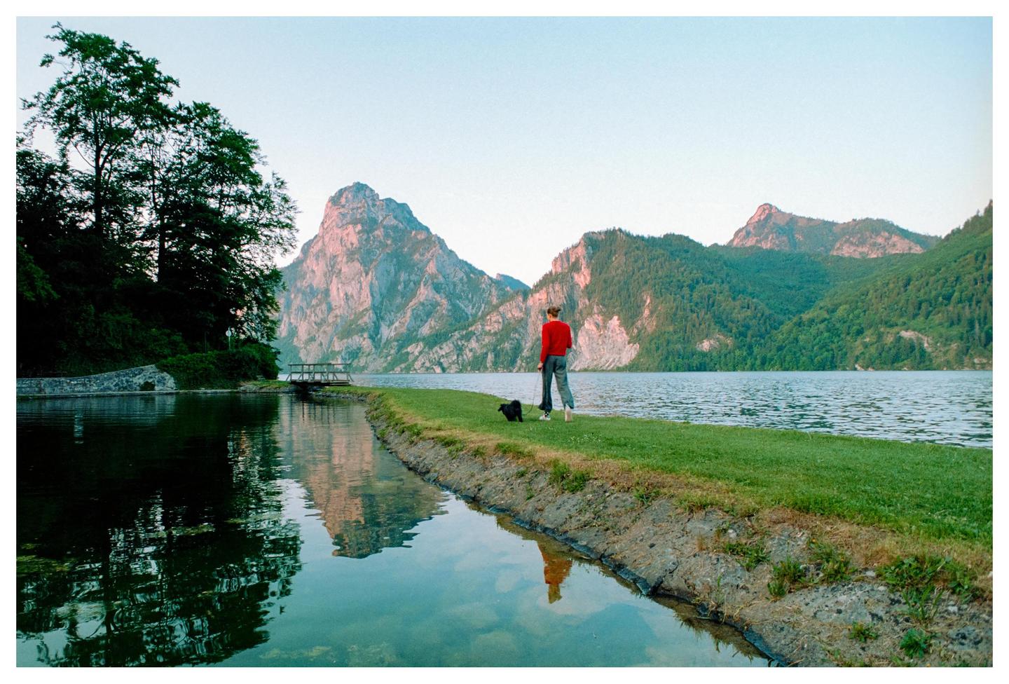 A woman in a red sweater walks her dog along a grassy bank next to a lake with mountains in the background.