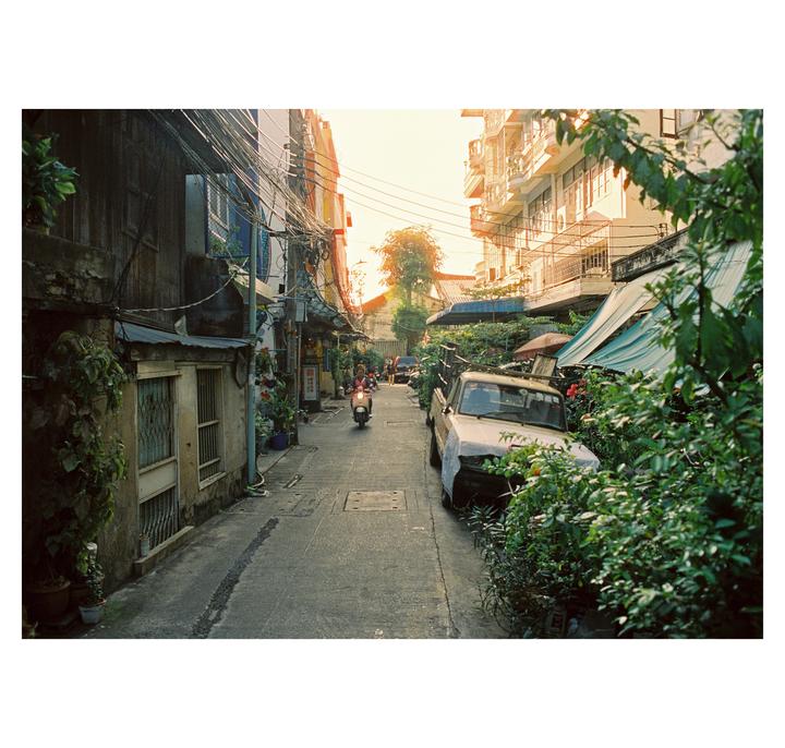 A narrow street in Bangkok is lined with buildings and lush greenery, with a scooter driving down the middle.