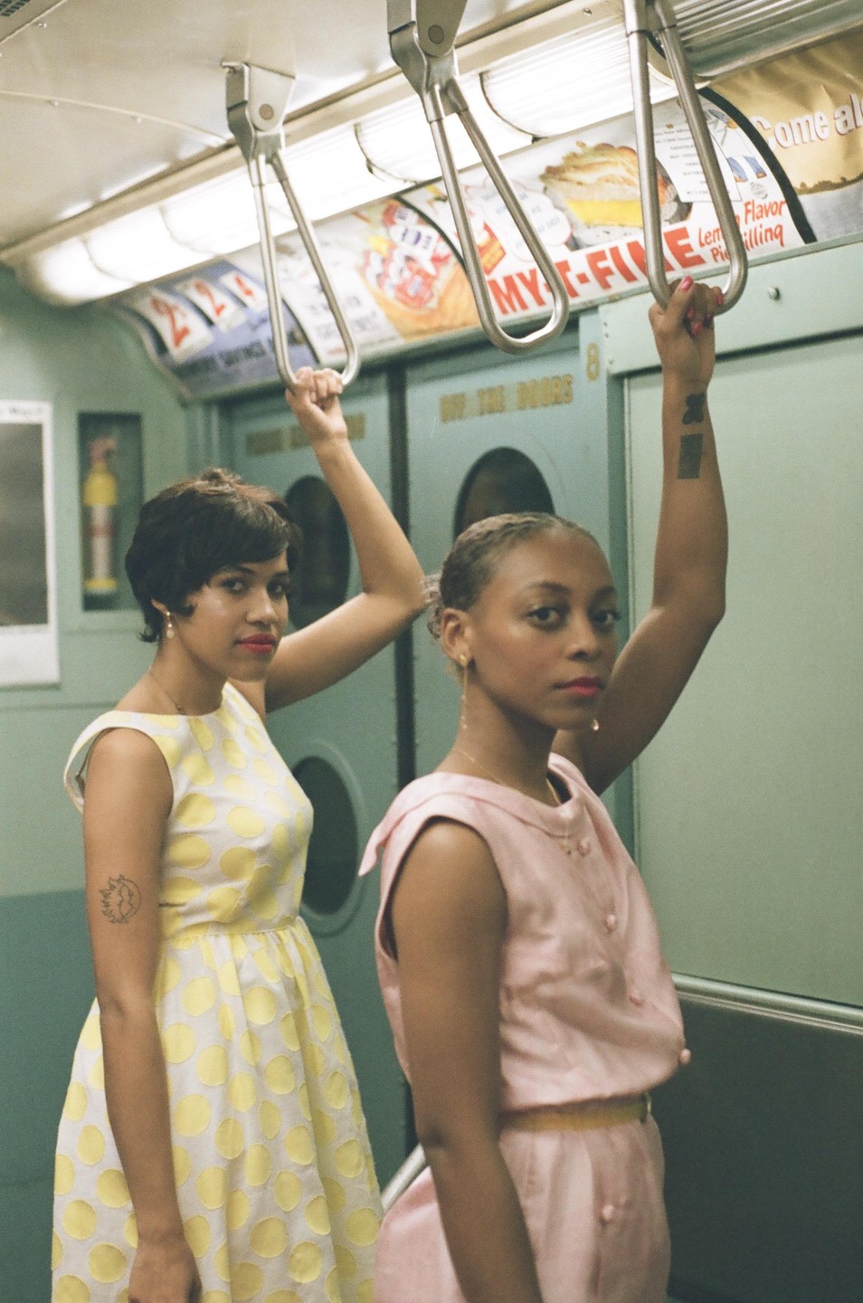 Two women in vintage dresses hold onto handrails inside a subway car.