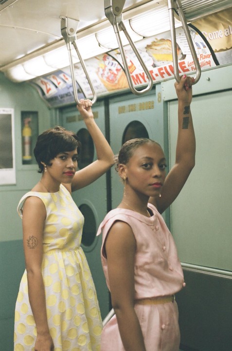 Two women in vintage dresses hold onto handrails inside a subway car.