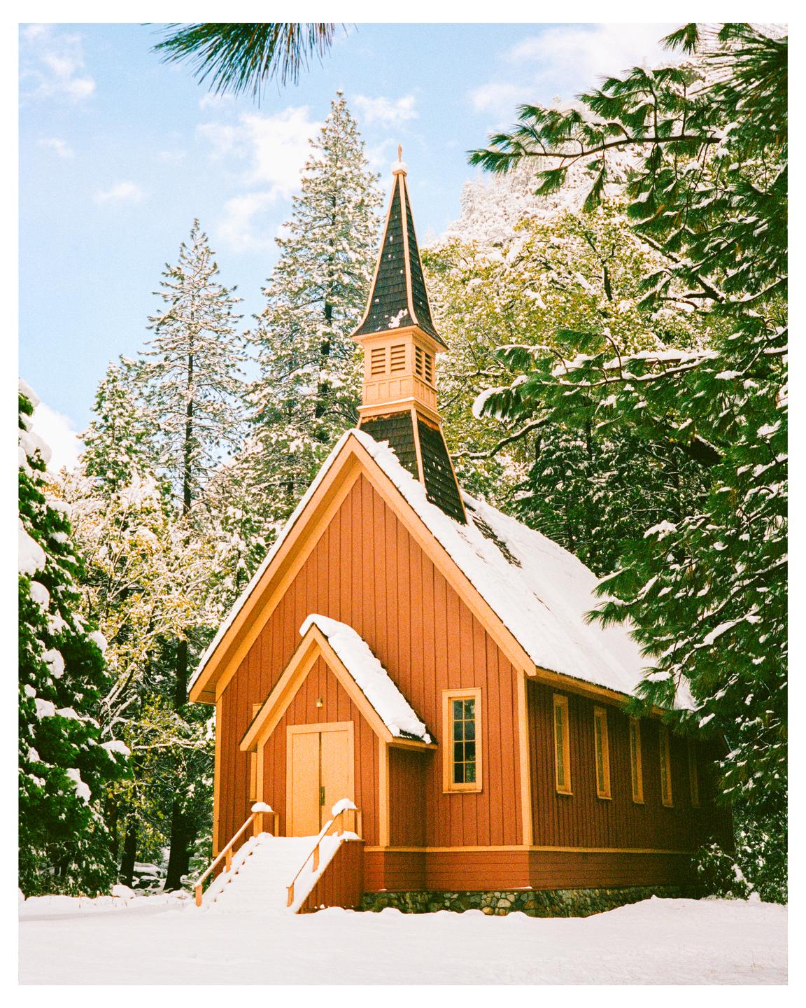 A small red chapel with a steeple is covered in snow, surrounded by snow-covered pine trees under a blue sky.