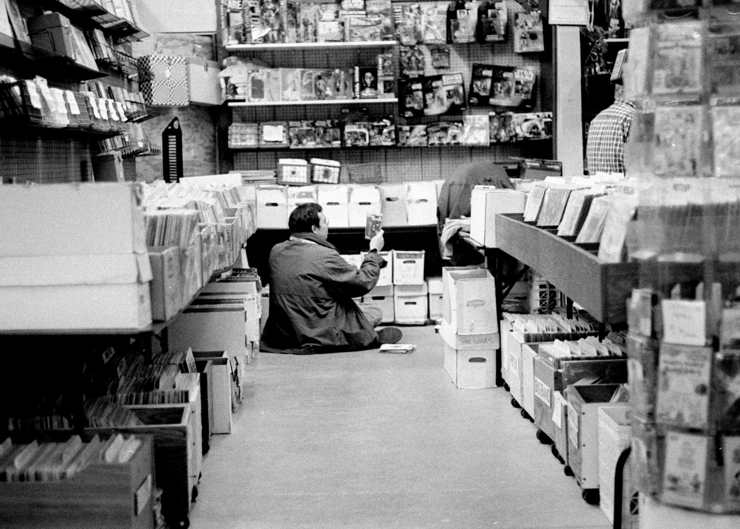 A man sits on the floor of a store, looking through items in a box.