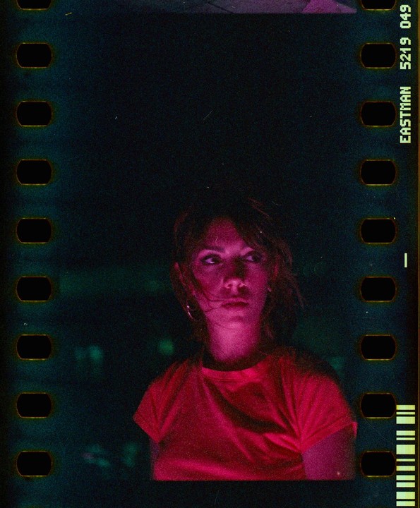 A woman with a serious expression is illuminated by red light in a dark setting.
