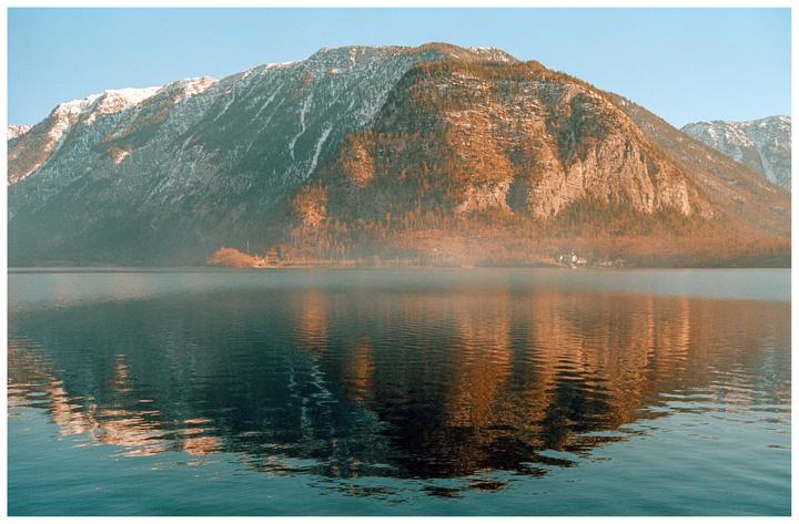 A large mountain with patches of snow and trees rises above a lake with a small village on its shore.