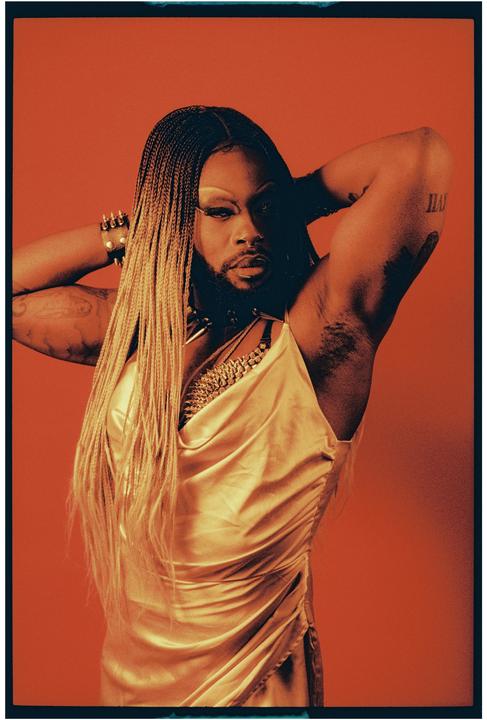 A person with long braided hair and a beard poses against a red background, wearing a gold dress and jewelry.
