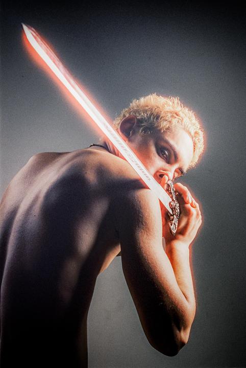 A man with short blond hair holds a glowing sword across his face, looking over his shoulder.