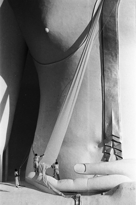 Three men are working on a giant Buddha statue, adjusting a large piece of cloth draped over its body and hand.