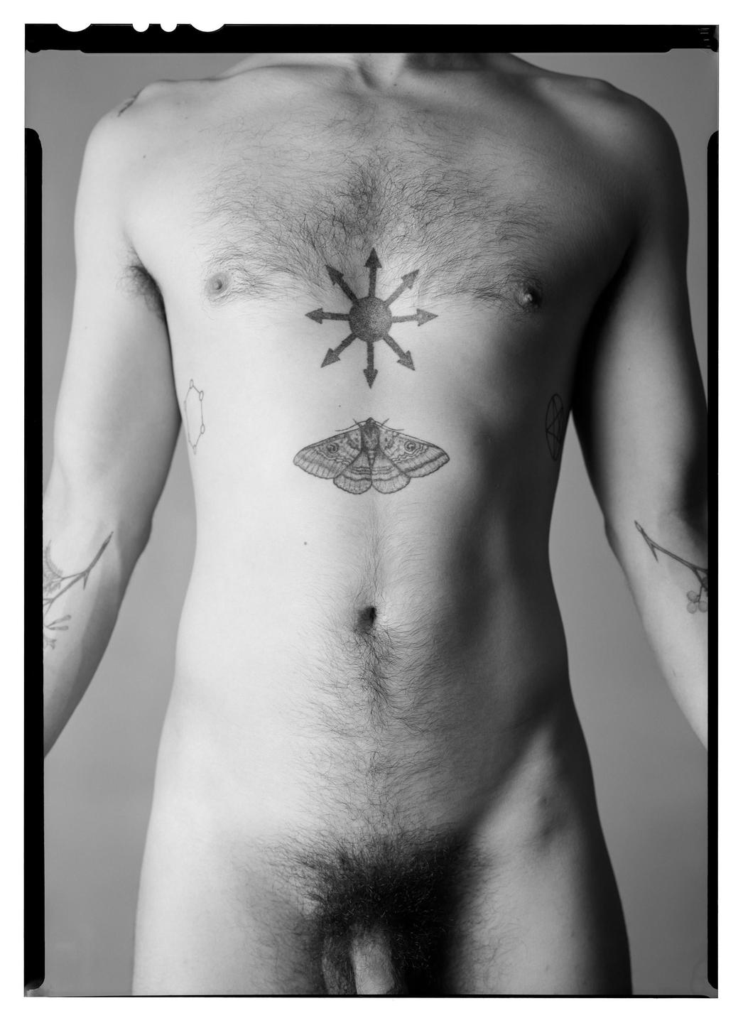 A man stands nude with tattoos on his chest and arms.