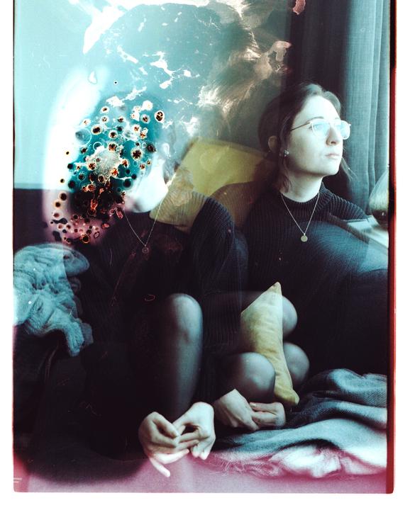 A woman with glasses sits looking out a window, with a double exposure effect and chemical damage on the left side of the image.