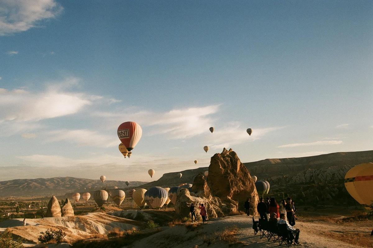 Hot air balloons ascend over a rocky, arid landscape with distant mountains under a partly cloudy sky.
